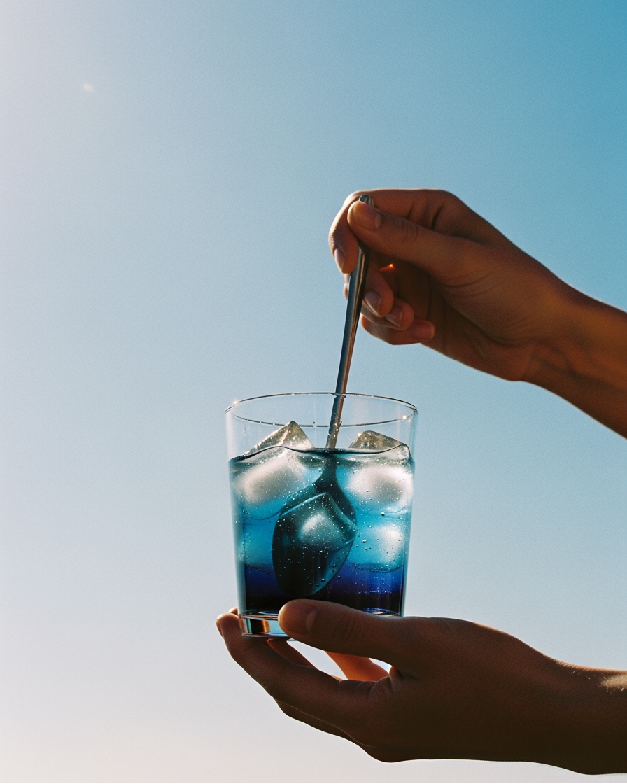 Person stirring a blue glass of methylene blue with water and ice against a clear blue sky. - USP Grade Methylene Blue